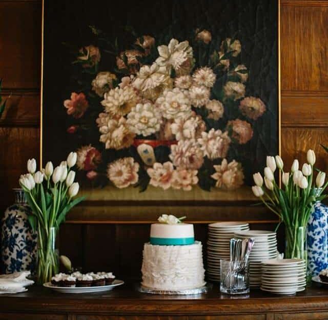 A wedding cake, plates, and forks sit on an antique table in front of a floral painting.