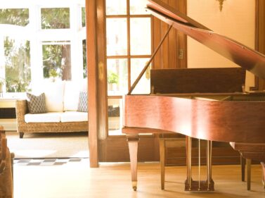 A wooden grand piano sits in a sunny room with a couch and sunroom in the background