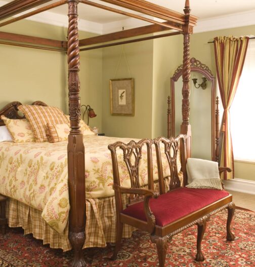 An elegant four post bed rests in a room with oriental rugs, antique furnishings, a large window, and light green walls.